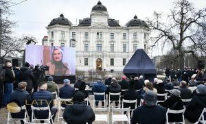 Lidija Lasmane ekrānā uzrunā atklāšanas sarīkojuma dalībniekus. Labā pusē vēl neatklātais piemineklis. FOTO Ilmārs Znotiņš - Valsts prezidenta kanceleja.